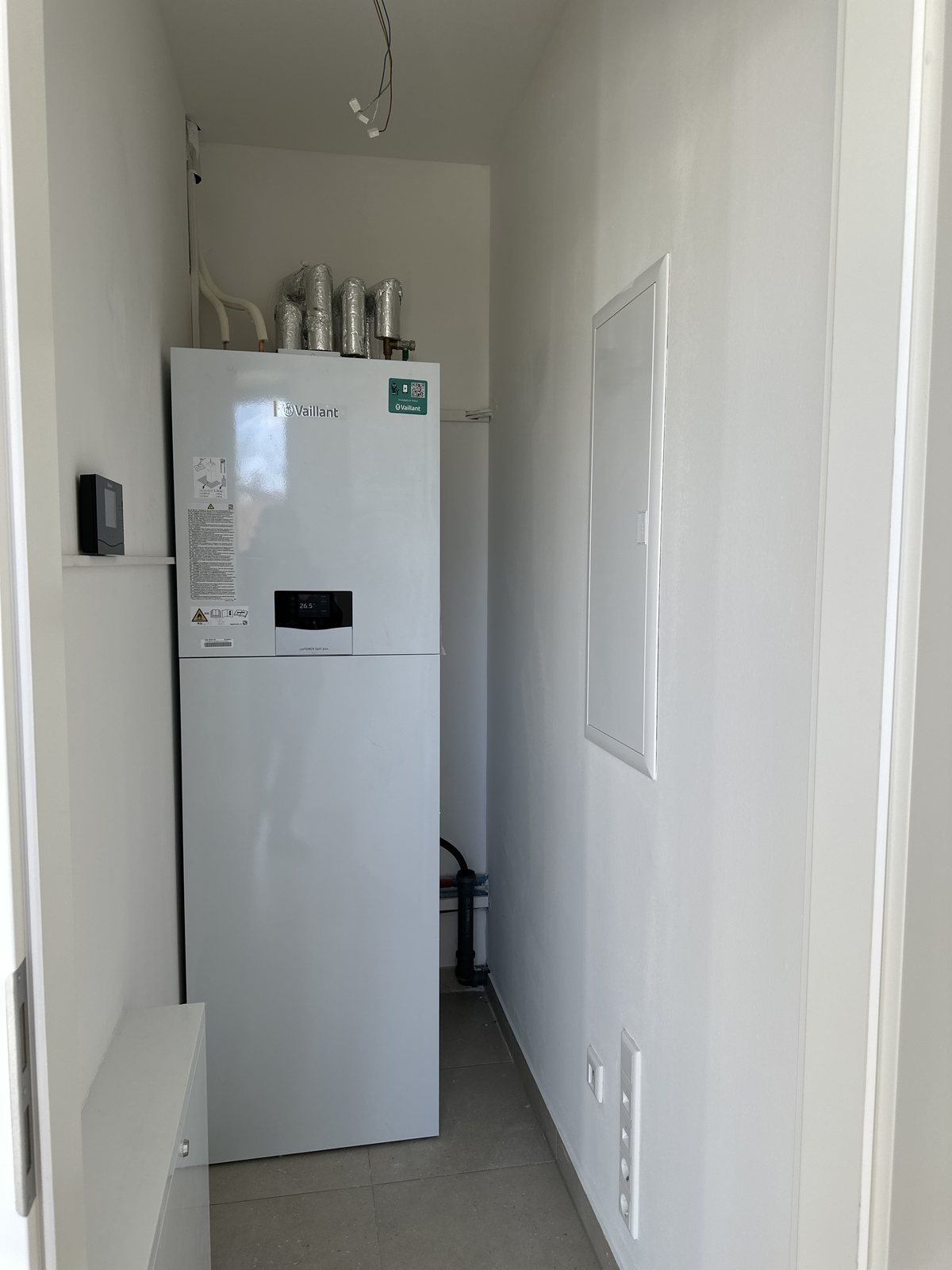 Weiße Wärmepumpen-Inneneinheit mit integriertem Speicher im Technikraum einer Wohnung, darüber gedämmte Anbindeleitungen