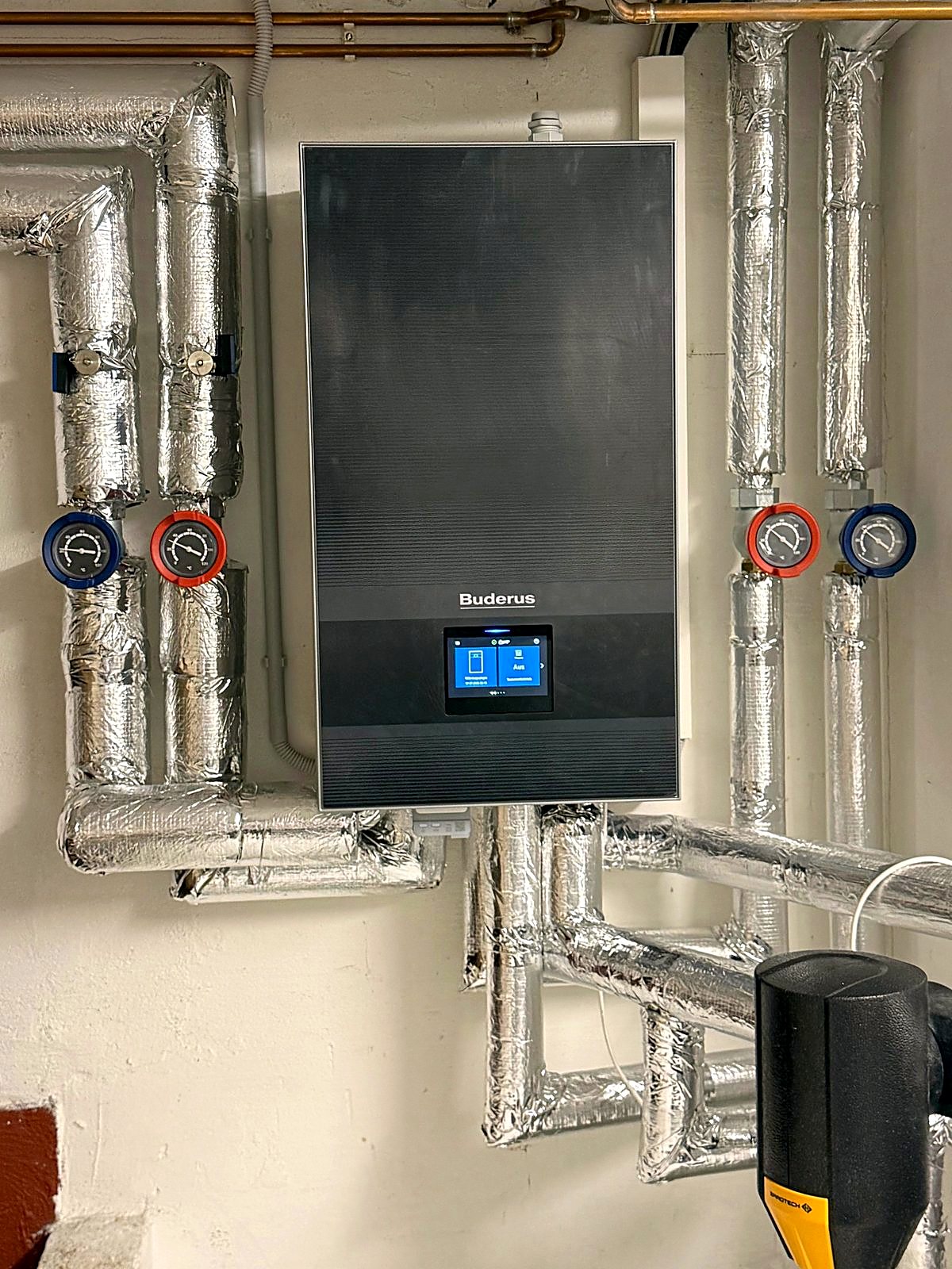 Buderus Wärmepumpen-Inneneinheit an der Wand mit gedämmten Rohrleitungen und Vorlauf- und Rücklaufthermometern