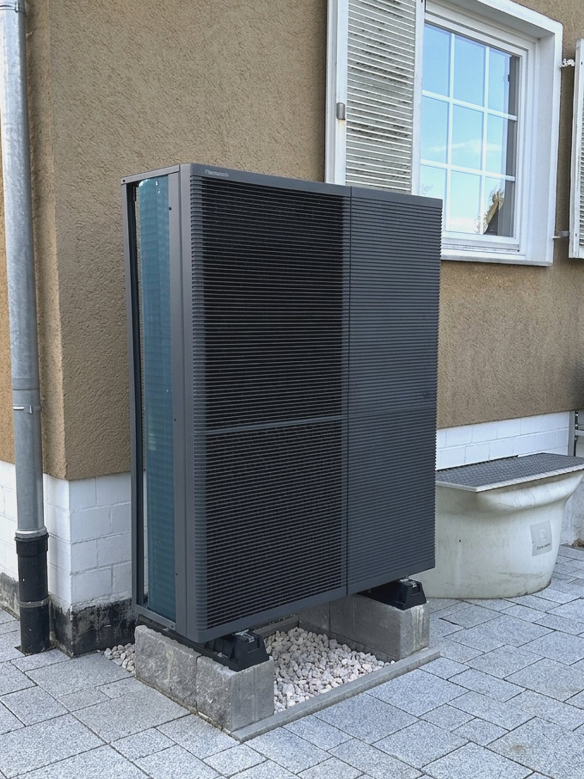 Wärmepumpen-Außengerät auf Schwellen an der Hauswand, mit Kiesbett und abgedichtetem Sockel